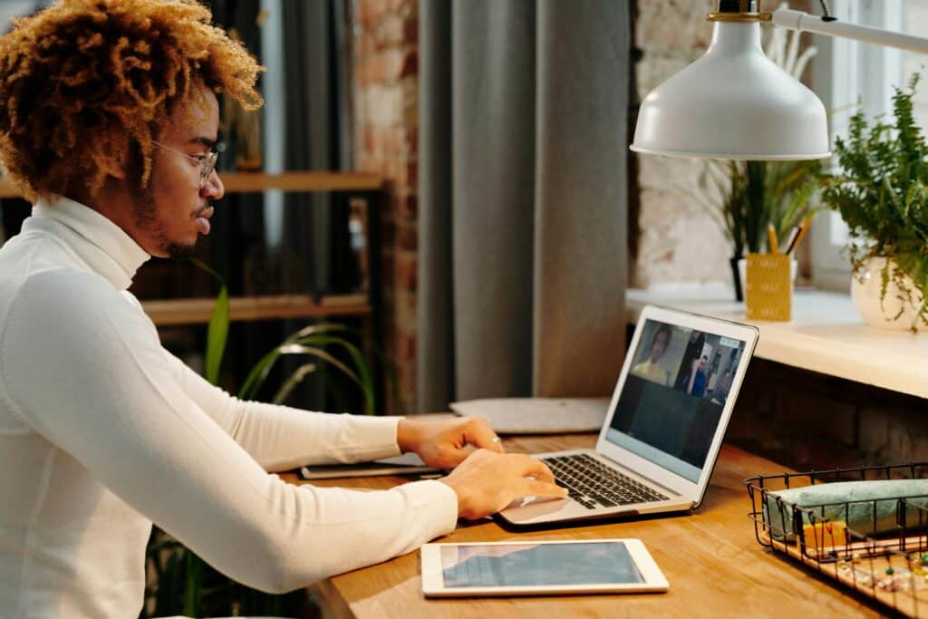 A person in a white turtleneck uses a laptop for a video call at a wooden desk with plants and a lamp in a cozy workspace.