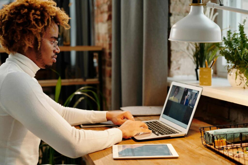 A person in a white turtleneck uses a laptop for a video call at a wooden desk with plants and a lamp in a cozy workspace.