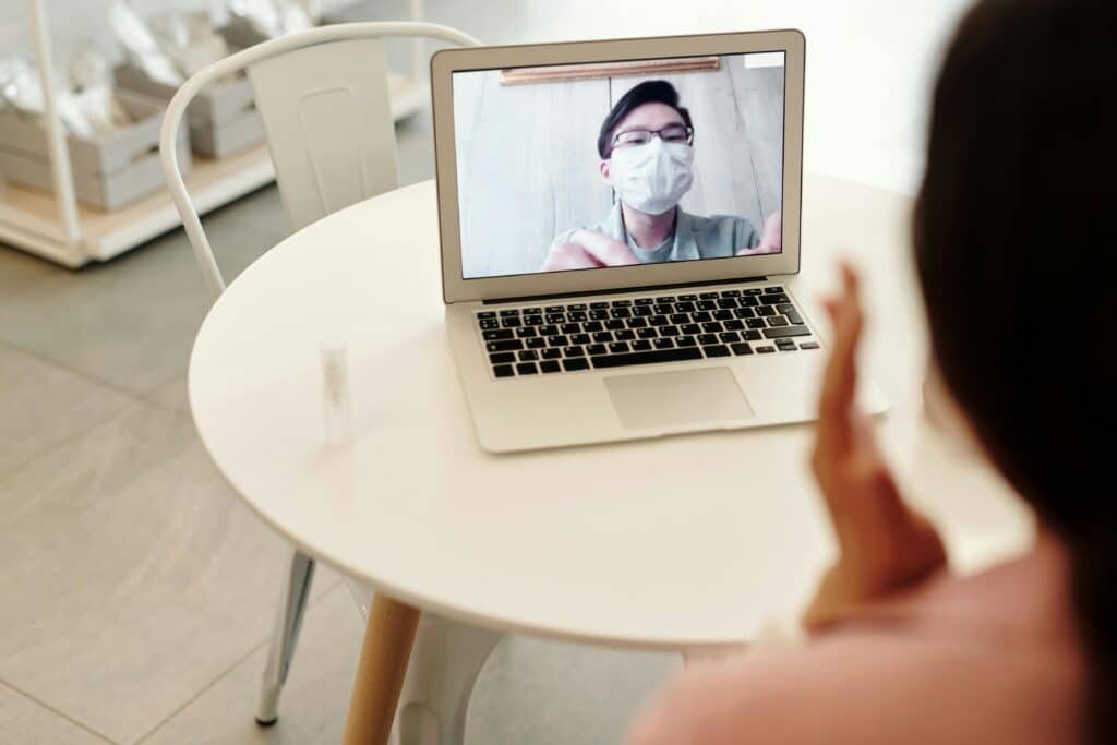A person waves while engaged in a video call on a laptop sitting on a round, white table in a modern, bright interior.