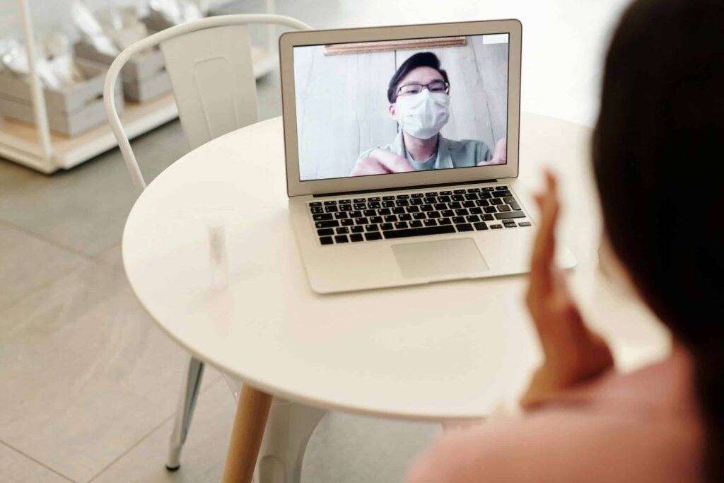A person waves while engaged in a video call on a laptop sitting on a round, white table in a modern, bright interior.
