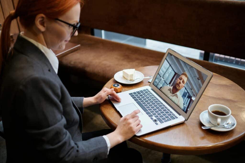 A woman in a suit with red hair is on a video call using a laptop at a café. A coffee and cake are on the table, creating a professional yet relaxed atmosphere.