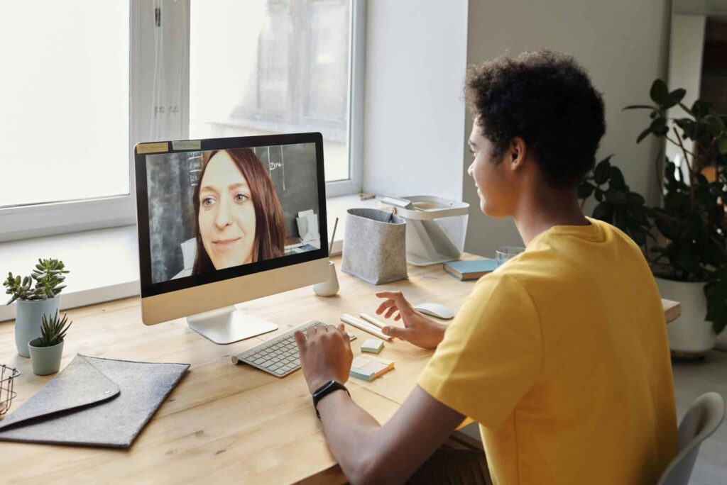A person in a yellow shirt interacts with a computer on a wooden desk, surrounded by plants and stationery, during a video call.
