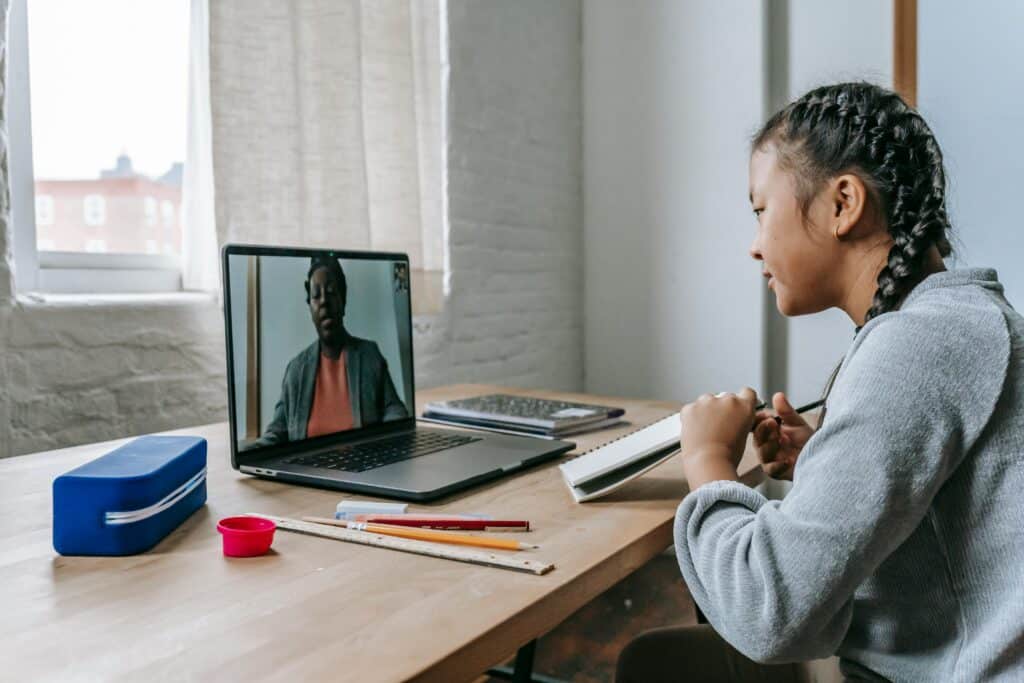 A young girl with braided hair sits at a desk, engaged in a virtual meeting on her laptop. Art supplies are scattered around her.