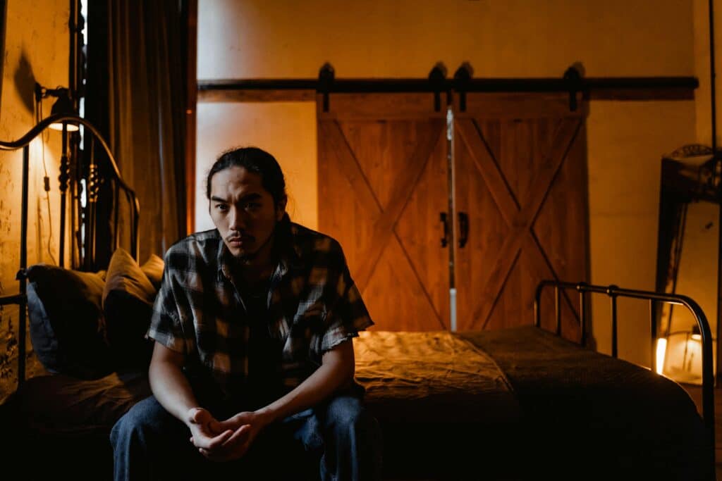 A person sits on a bed in a warmly lit room, appearing contemplative amidst rustic decor and wooden sliding doors.
