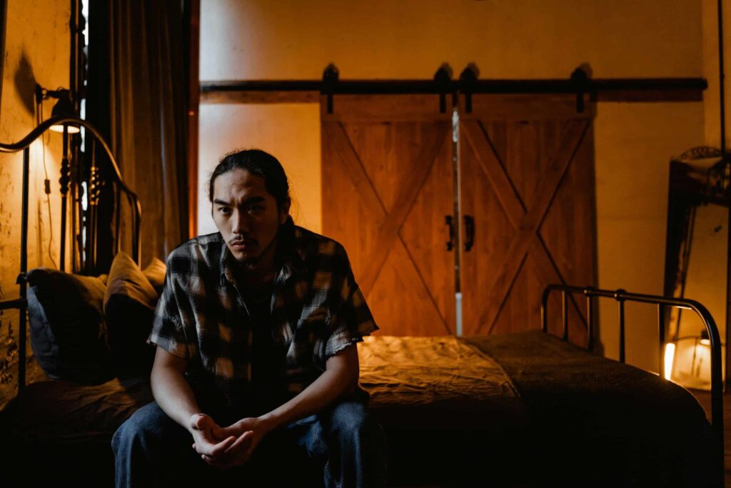 A person sits on a bed in a warmly lit room, appearing contemplative amidst rustic decor and wooden sliding doors.