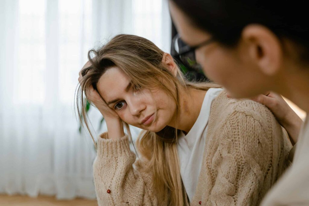 A woman sits with her head in her hands, visibly distressed, while another person comforts her from the side in a softly lit room.