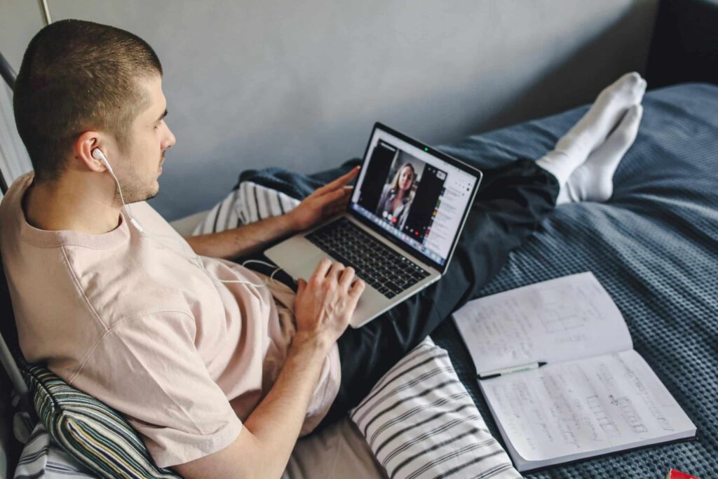 A person sits on a bed, wearing headphones and using a laptop for a video call, with notebooks and papers beside them.