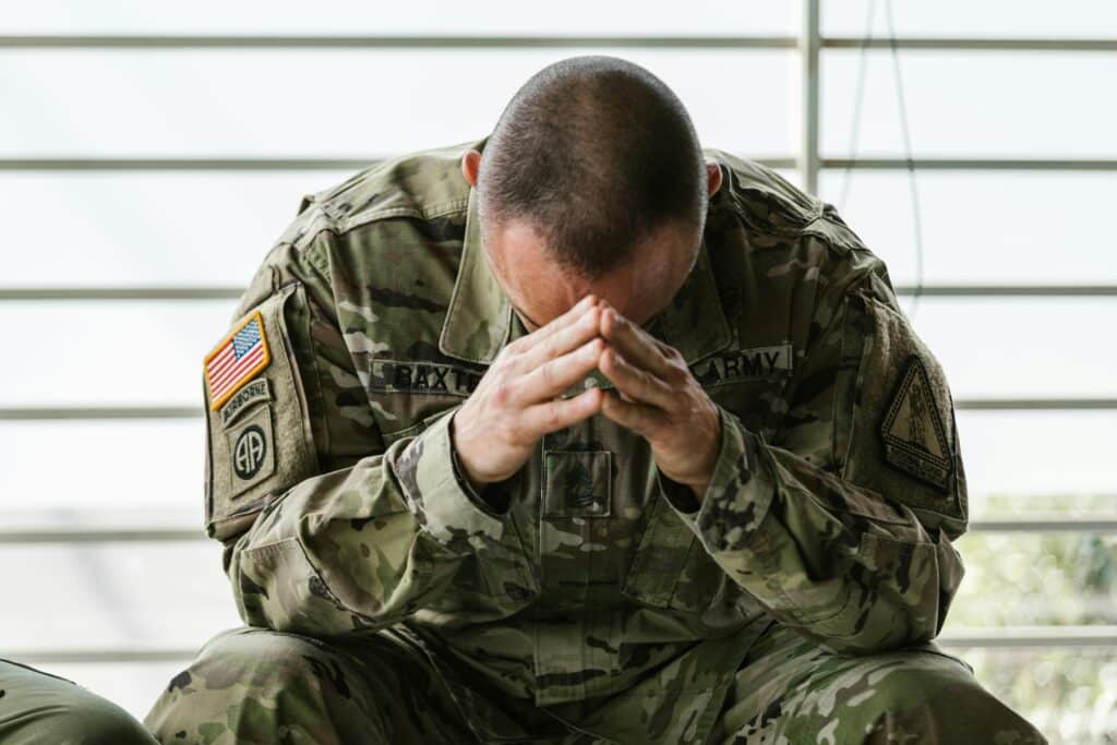 A soldier in military uniform sits with his head bowed and hands clasped, reflecting a moment of deep contemplation or distress.