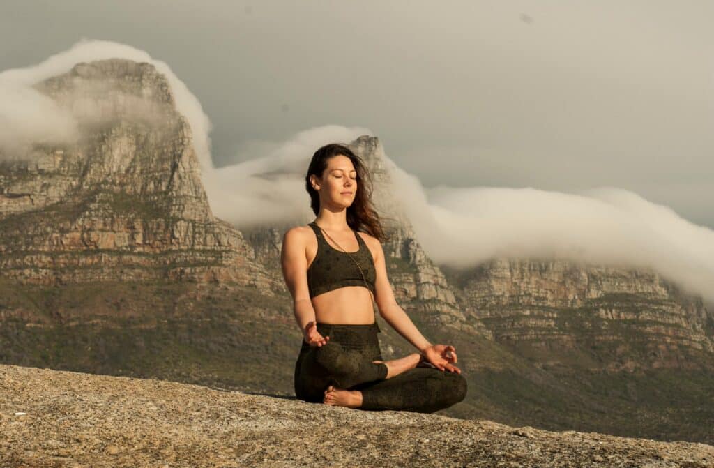 A person in yoga attire meditates peacefully on a rocky ledge with majestic mountains and soft clouds in the background.