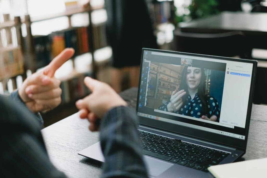 A person gestures animatedly while participating in a video call on a laptop, with a bookshelf in the background.