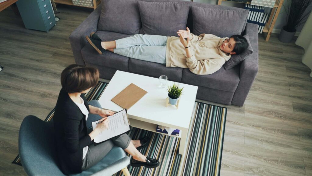 A therapist sits with notes as a patient relaxes on a couch during a counseling session in a cozy, modern office.