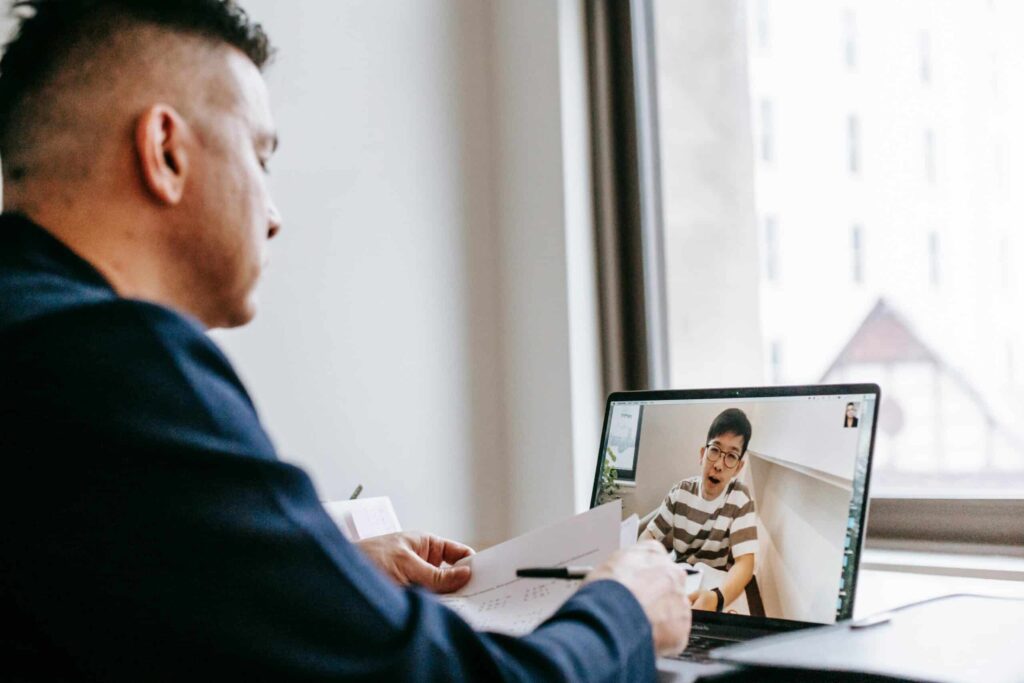 A person sits at a desk, participating in a video call on a laptop, displaying a participant in a striped shirt on screen.