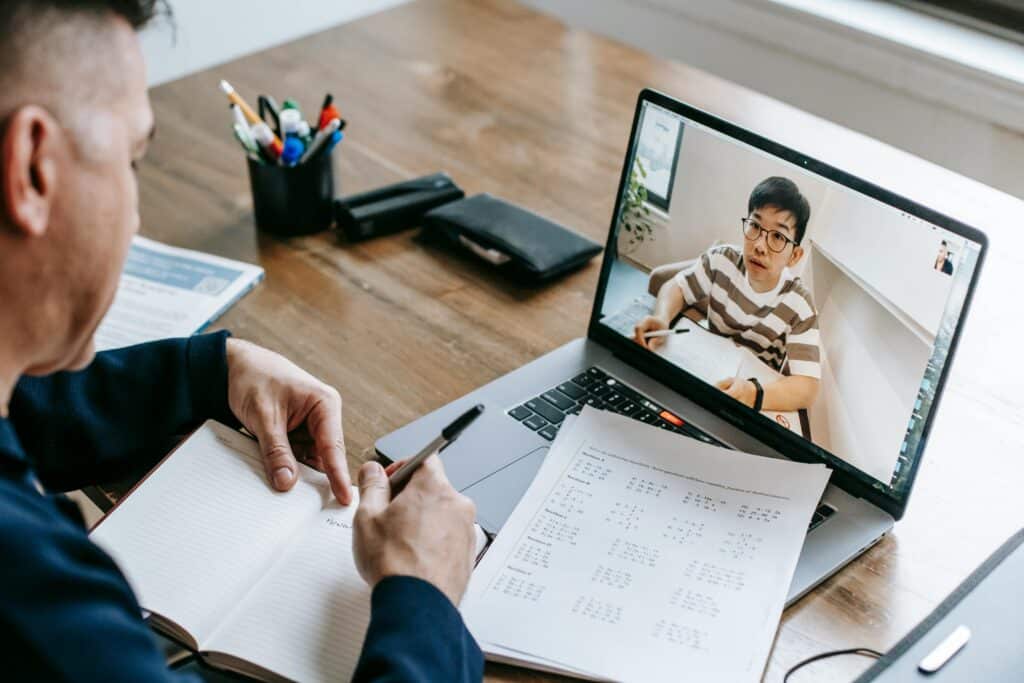 A person sits at a desk with a notebook and pen, engaged in a virtual learning session on a laptop.