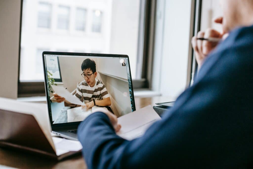 A person participates in a video call on a laptop, reviewing documents while seated at a desk near a window.