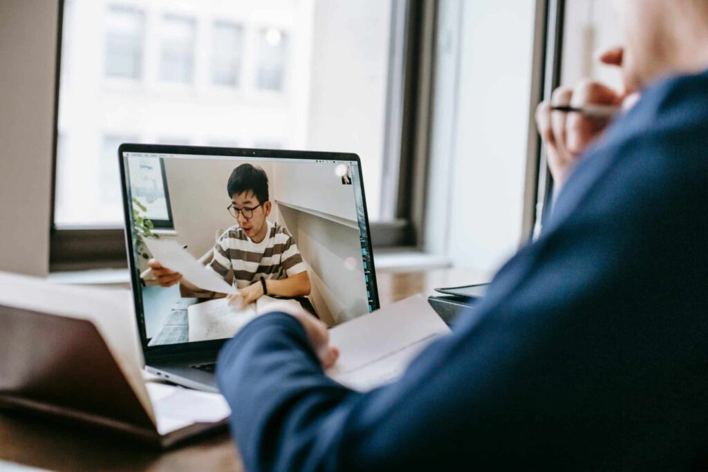A person participates in a video call on a laptop, reviewing documents while seated at a desk near a window.