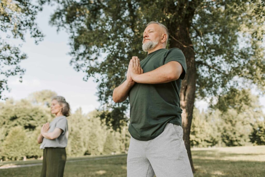 Two individuals practice yoga outdoors, standing serenely in a park surrounded by trees on a sunny day.
