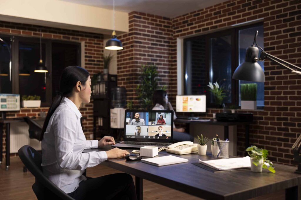 A woman in a white shirt participates in a video call on a laptop in a modern office with brick walls and plants, conveying a professional tone.