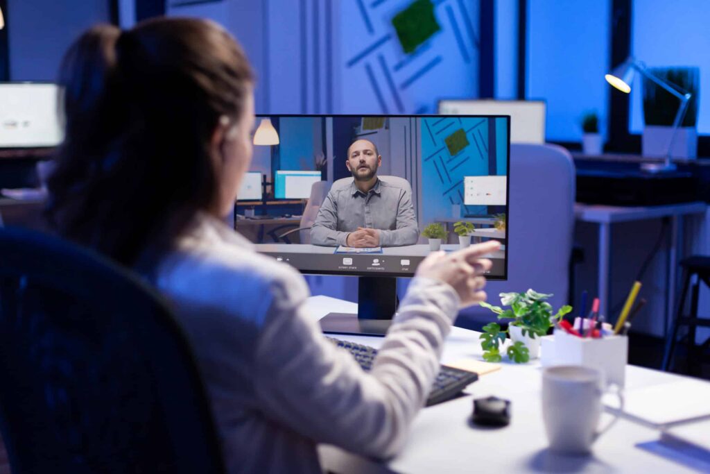 A woman in an office video chats on a computer screen. She points at a man speaking. The room is softly lit with a blue hue, creating a focused atmosphere.