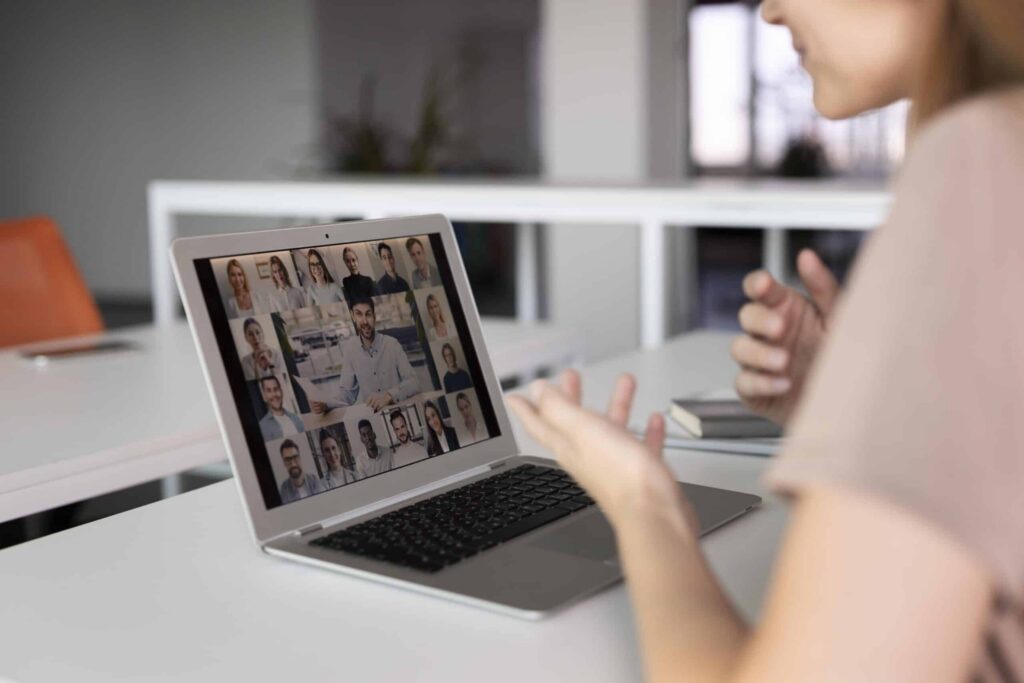A woman participates in a video conference on a laptop, displaying a grid of people in a virtual meeting. She's gesturing with her hands, indicating engagement.