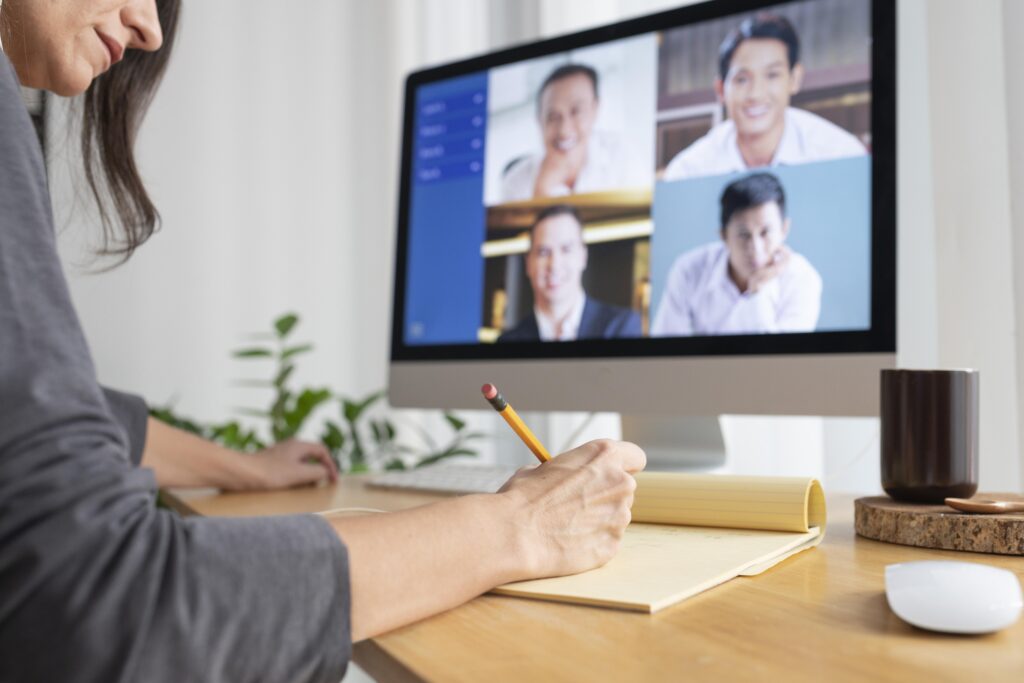 A person writes notes at a desk during a video conference, visible on a computer screen showing four smiling participants. The scene is calm and focused.