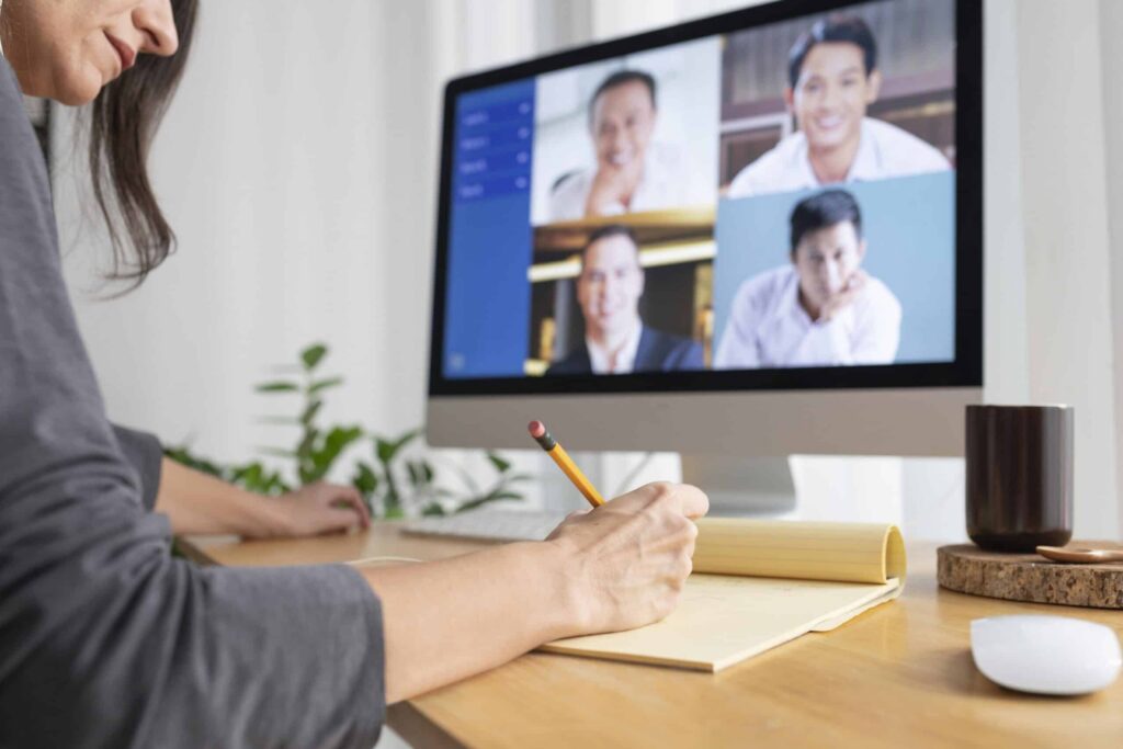 A person writes notes at a desk during a video conference, visible on a computer screen showing four smiling participants. The scene is calm and focused.