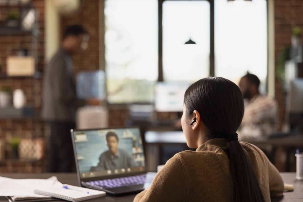 A woman with earbuds sits at a desk, focused on a video call on her laptop. A blurred office background features colleagues and a brick wall.