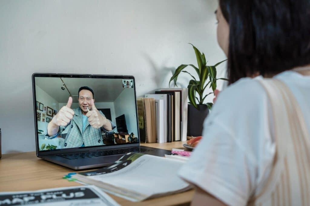 A person engages in a video call, giving a thumbs-up, while seated at a desk with books and plants in the background.