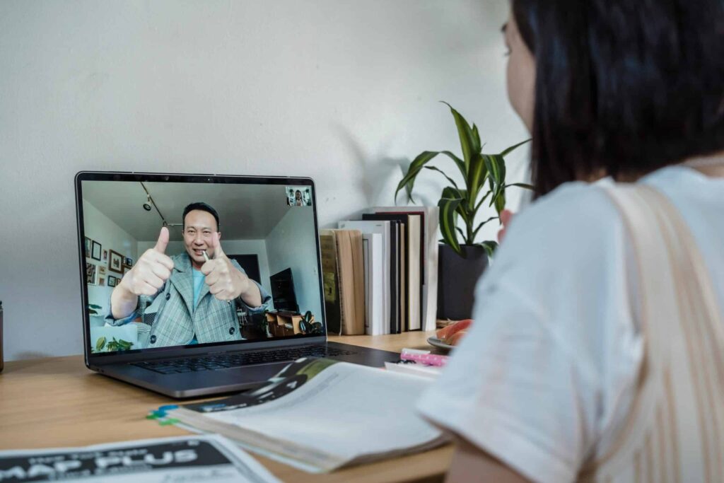 A person on a video call shows thumbs up on a laptop screen, while another individual looks on, surrounded by books and plants.