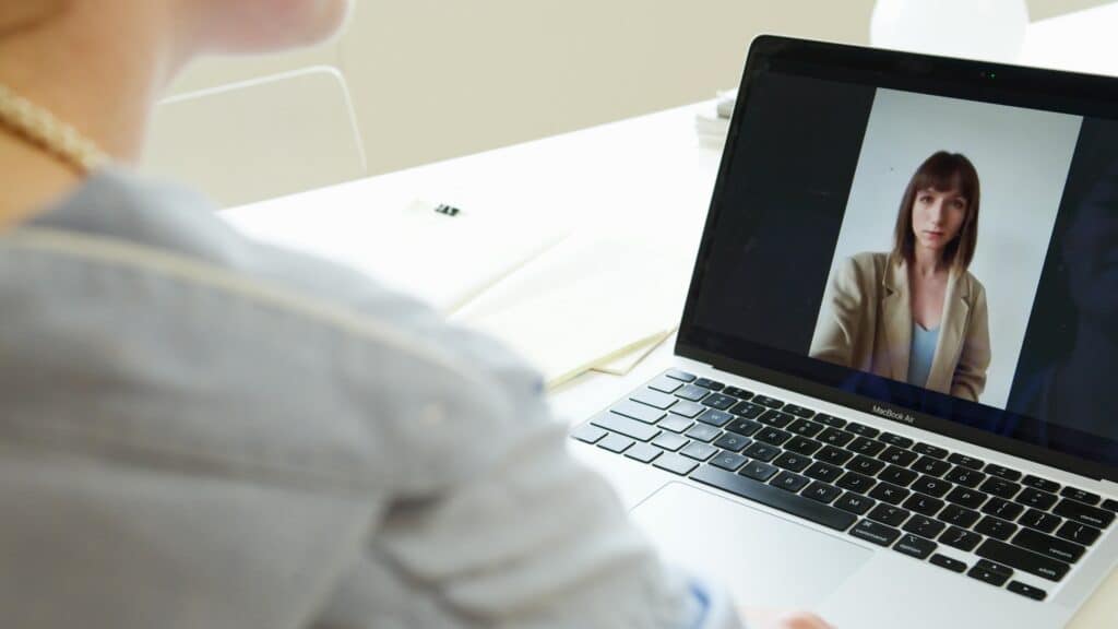 A person in a blue shirt participates in a video call on a laptop. The screen shows a woman in a beige blazer. The setting is a well-lit room.