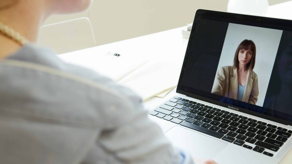 A person in a blue shirt participates in a video call on a laptop. The screen shows a woman in a beige blazer. The setting is a well-lit room.