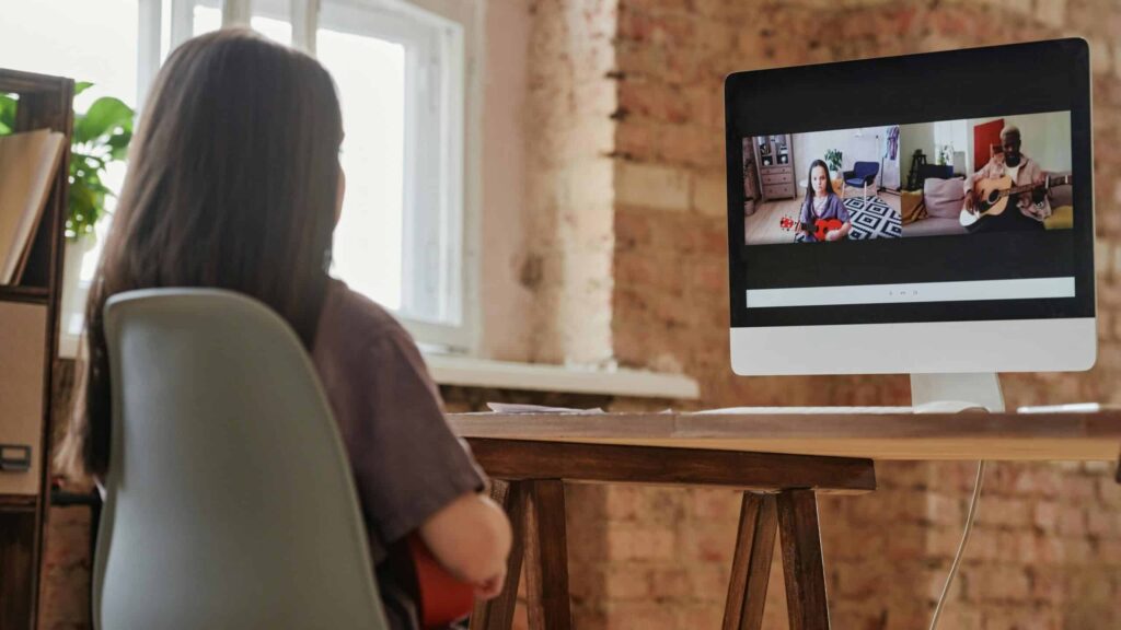 A person sitting at a desk with a computer, participating in a video call with two others, one playing a guitar, the other with a ukulele.