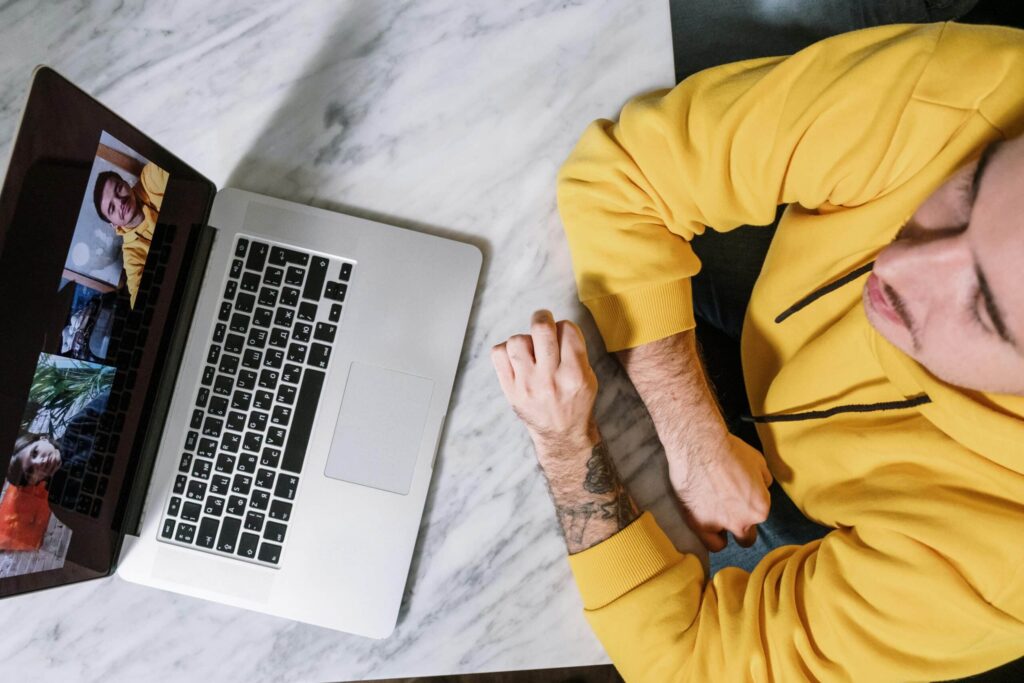 A person wearing a yellow hoodie sits at a marble table, engaged in a video call on a laptop displaying multiple faces.