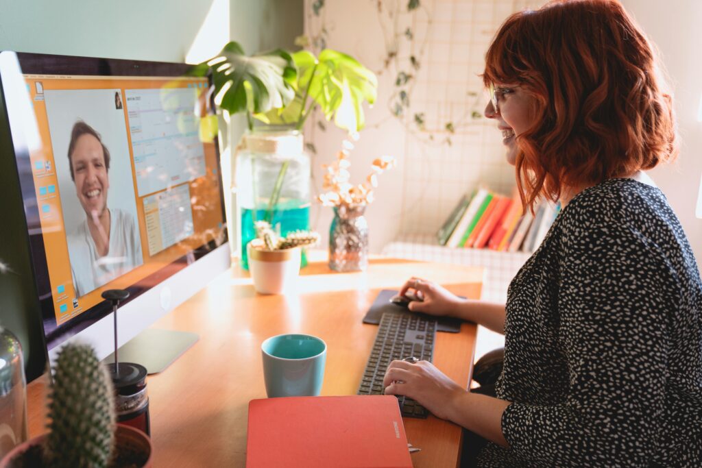 A woman with red hair smiles while video chatting on a computer. Her desk has a blue mug, notebook, and plants, creating a warm, friendly atmosphere.