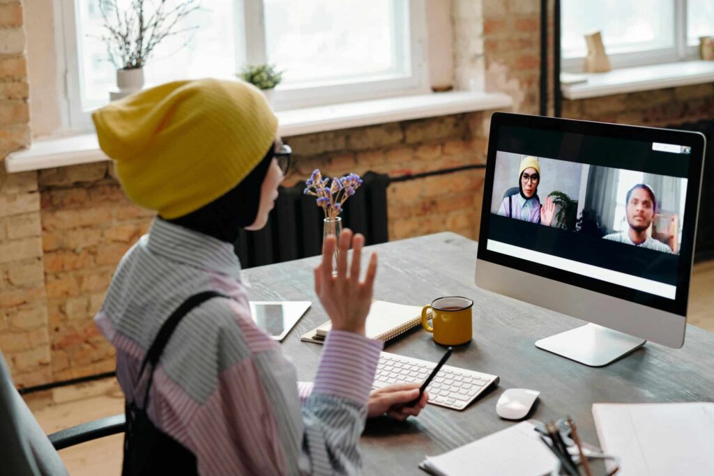 Person in a yellow hat waves at a computer screen showing two individuals on a video call. The room is bright with natural light, conveying a casual work setting.