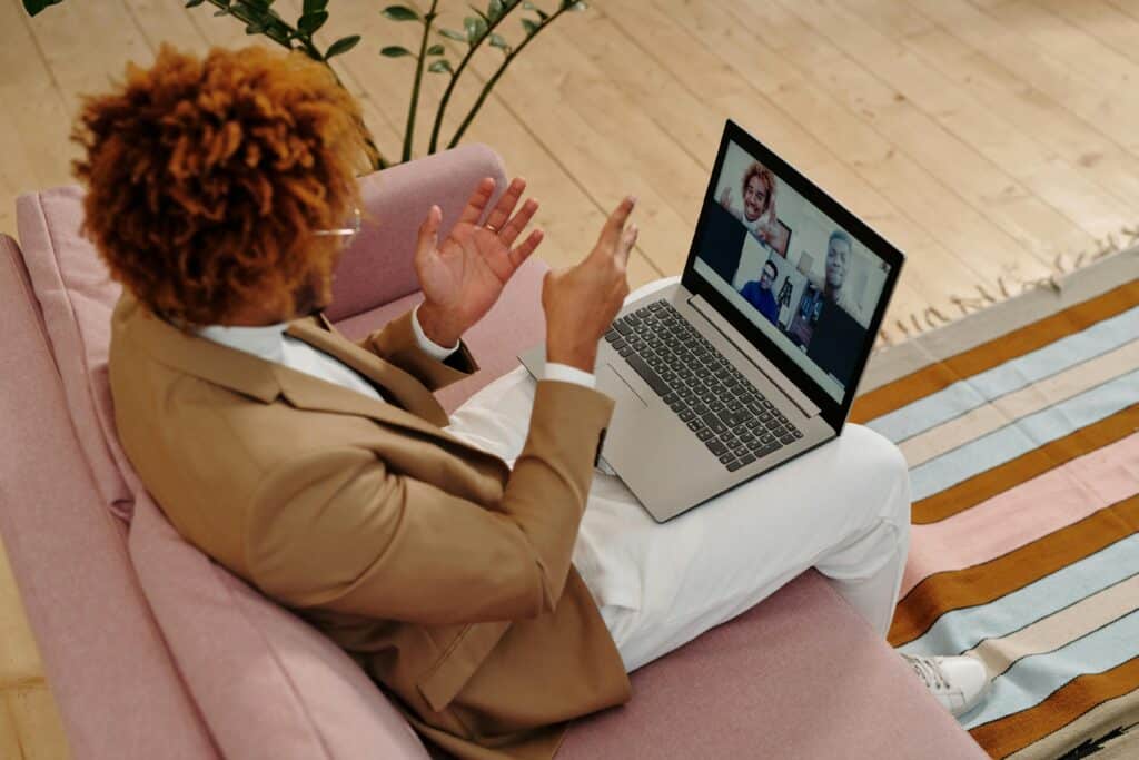 A person in a tan suit sits on a pink sofa, gesturing at a laptop displaying a video call with four people, conveying a professional virtual meeting.