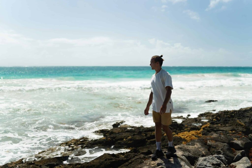 A person in a white shirt and khaki shorts stands on rocky shore, gazing at the turquoise sea and waves under a clear blue sky, conveying tranquility.