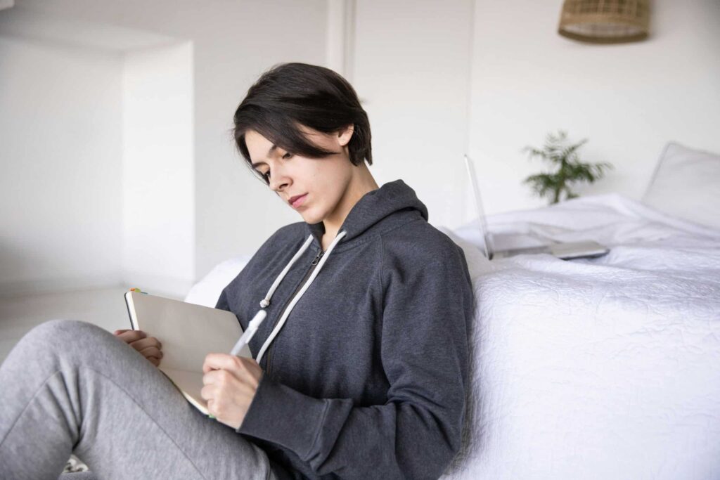 Young woman with short dark hair sits by a bed, writing in a notebook. She wears a gray hoodie and sweatpants, conveying a relaxed and focused mood.