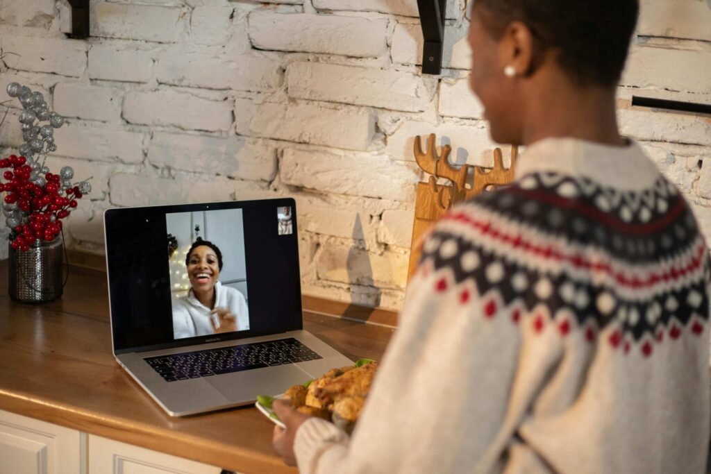 A person in a cozy sweater holds a plate of food in front of a laptop displaying a video call, with festive decor in the background.