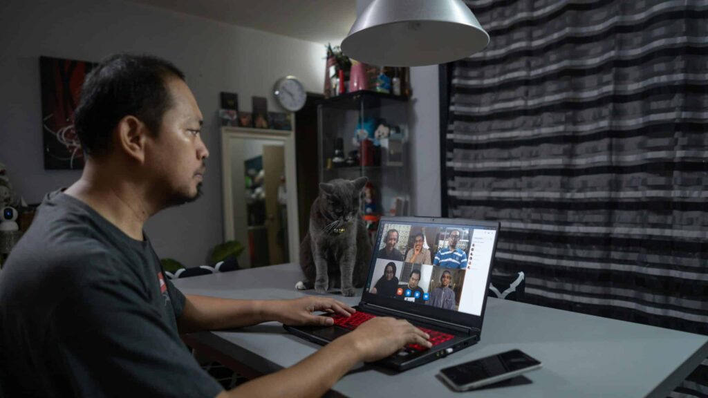A man works on a laptop at a table, attending a video conference. A gray cat sits beside the laptop. The room is dimly lit with a modern decor.