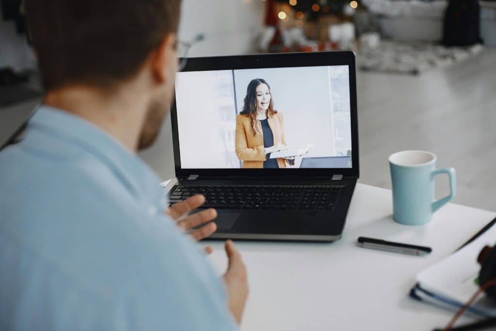 A man in a blue shirt sits at a table, participating in a video call on a laptop with a woman speaking. A blue mug and notebook are nearby, conveying a professional tone.