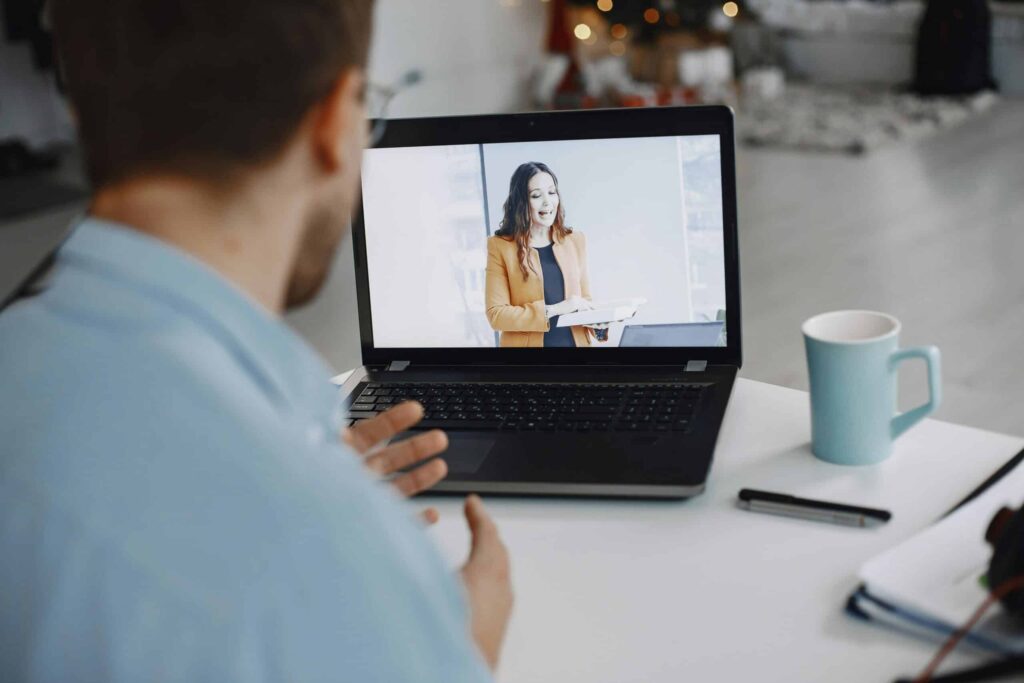 A person is having a virtual meeting, watching a woman on a laptop screen. The desk has a blue mug, pen, and papers, conveying a professional setting.