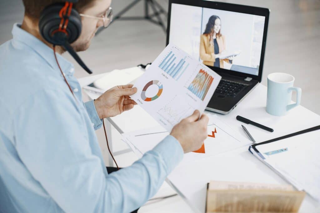 A man wearing headphones reviews data charts in a bright office setting. A woman in business attire appears on a laptop screen, holding a document.
