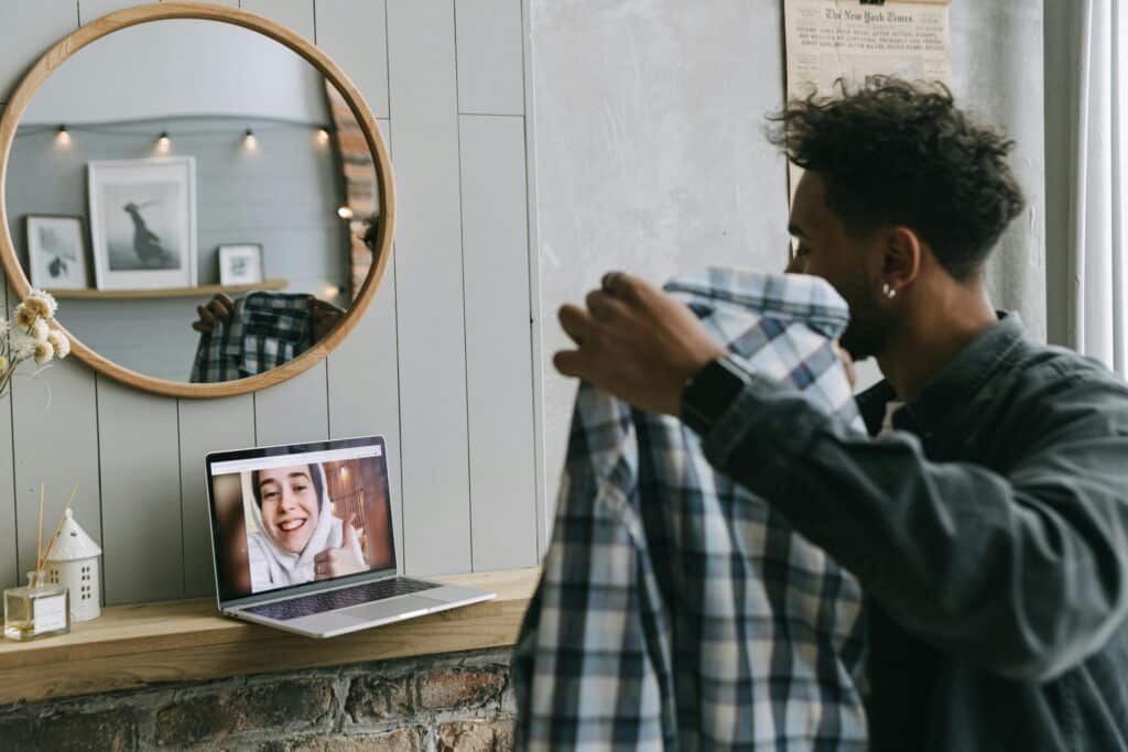A man holds a plaid shirt, facing an open laptop with a smiling woman on a video call. A round mirror and soft, ambient lighting set a cozy tone.