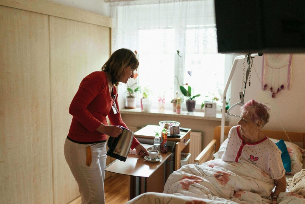 A caregiver in a red shirt prepares a cup of tea by a bedside table in a cozy, sunlit room with plants and personal items.