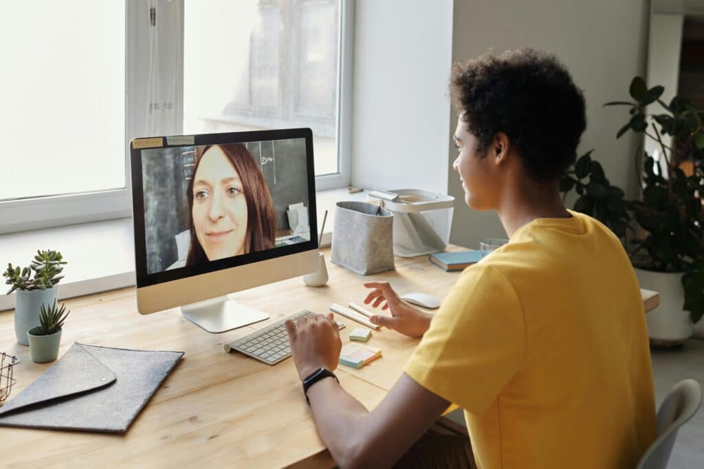 A person in a yellow shirt participates in a video call at a desk, viewed in profile. A woman's face is on the computer screen. The setting is bright and organized.