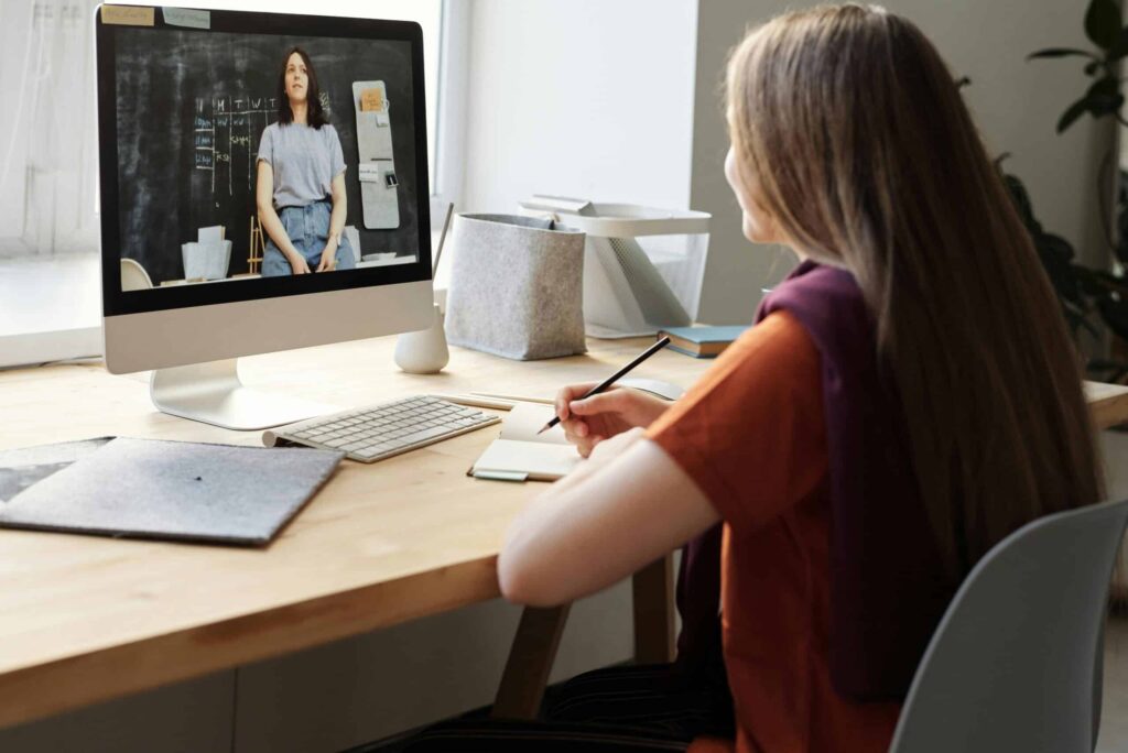 A woman sits at a desk, engaging in a video call on her computer. She takes notes while attentively watching a teacher on the screen with a chalkboard.