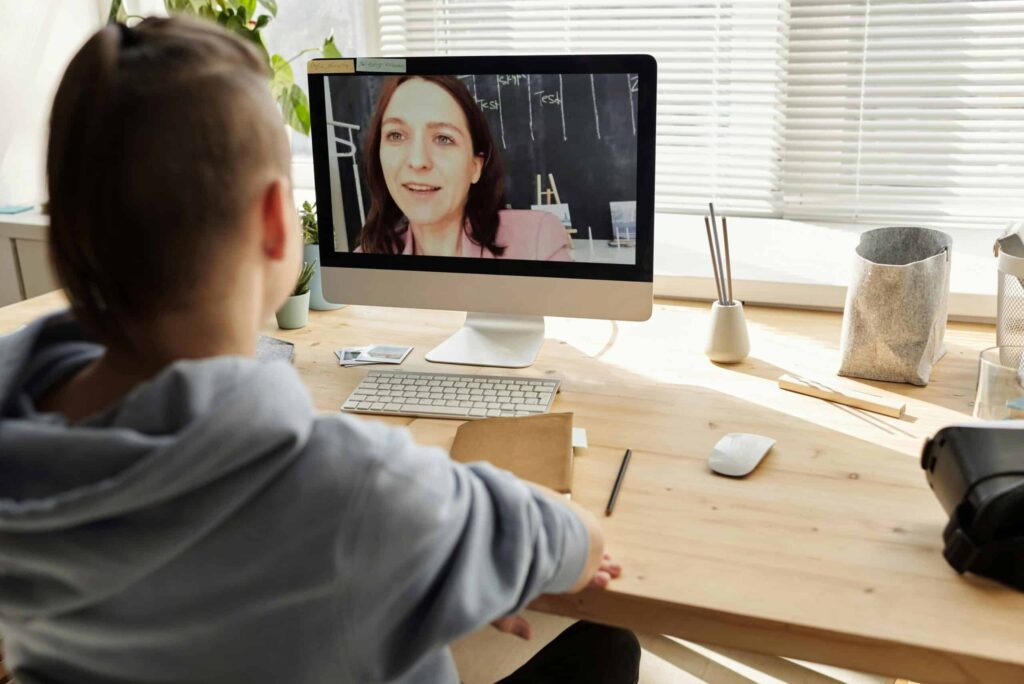 A person in a hoodie is sitting at a wooden desk, engaged in a video call on a computer. A woman on the screen appears friendly and professional. The setting is a bright room with natural light.