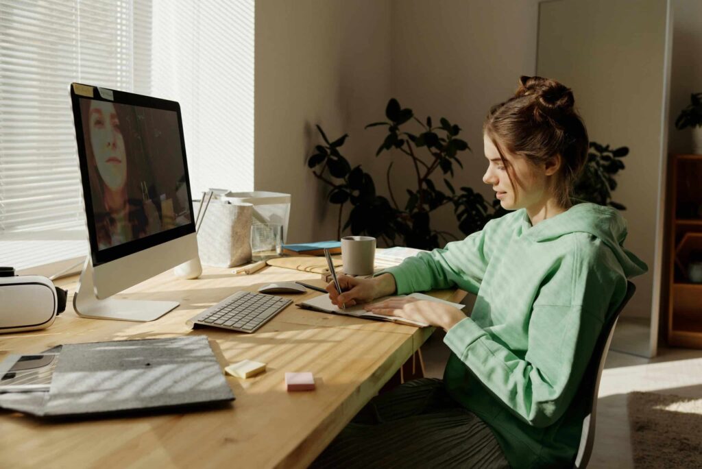 A woman in a green hoodie sits at a desk, writing in a notebook during a video call on a computer. Sunlight filters in, creating a calm atmosphere.