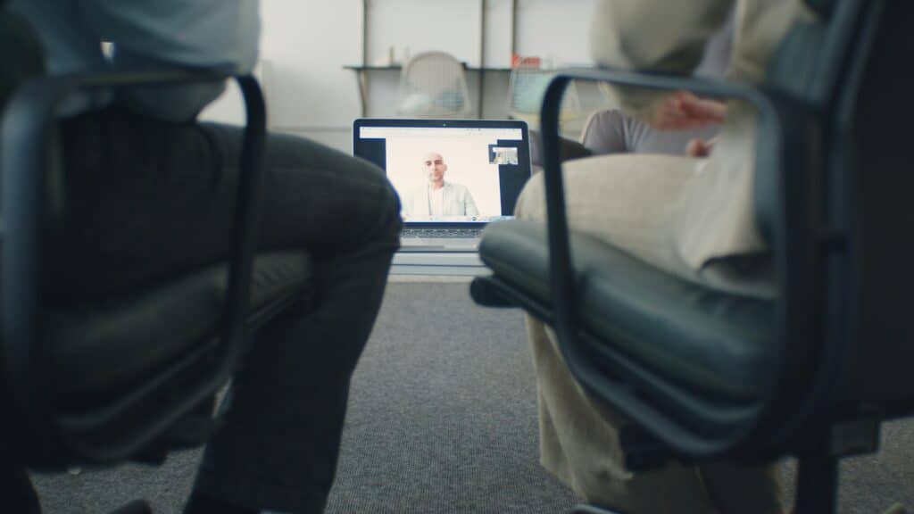 Two people sit in office chairs facing a laptop on a table, participating in a video call with a person displayed on the screen, creating a professional tone.