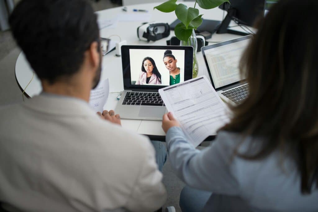 Two individuals are in a video call on a laptop, while another person holds a document at a modern workspace with plants.